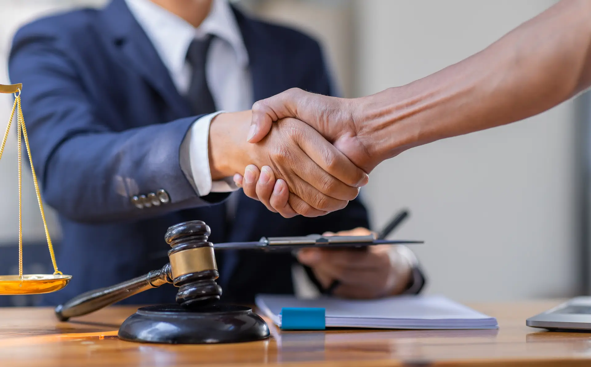 Close up shot of client shaking lawyers hand over desk that has gavel, scales of justice, and paperwork on it.