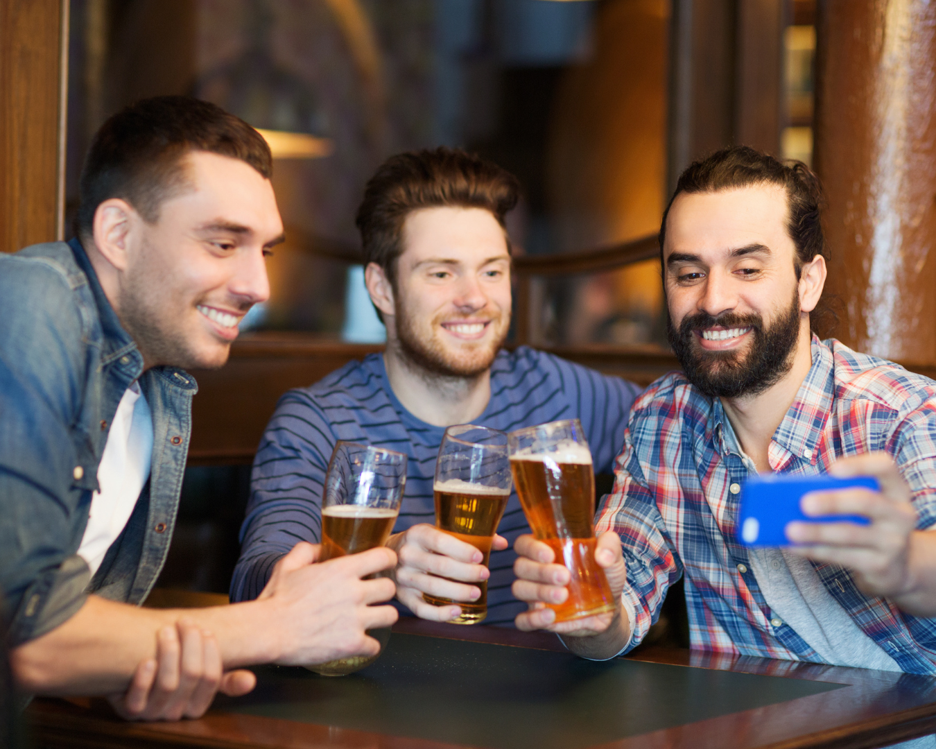 Three adult men, sitting at a table in a bar, with beers in hand, taking a selfie.