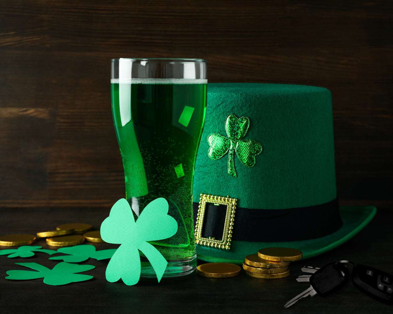 St. Patrick's Day hat, four leaf clovers, gold coins, car keys, and pint of green beer on wooden table.